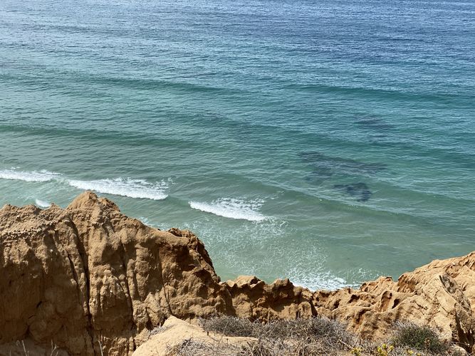The image shows a coastal cliff with layers of sedimentary rock in various shades of beige and brown. At the base, there's a rocky shoreline with flat rock formations extending into the water, where waves in blue and green tones gently wash over them.